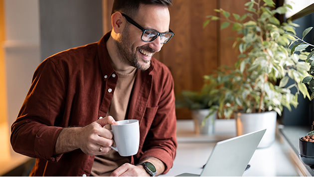 Casual, happy man with coffee cup looking at his laptop in an open and airy workspace